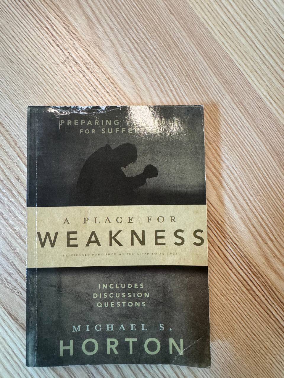 机の上に置かれた『A Place for Weakness』のペーパーバック。上部に「PREPARING YOURSELF FOR SUFFERING」、中央の帯に大きく「A PLACE FOR WEAKNESS」、下に著者名 MICHAEL S. HORTON。表紙には膝をついて祈る人のシルエットが暗く写っている