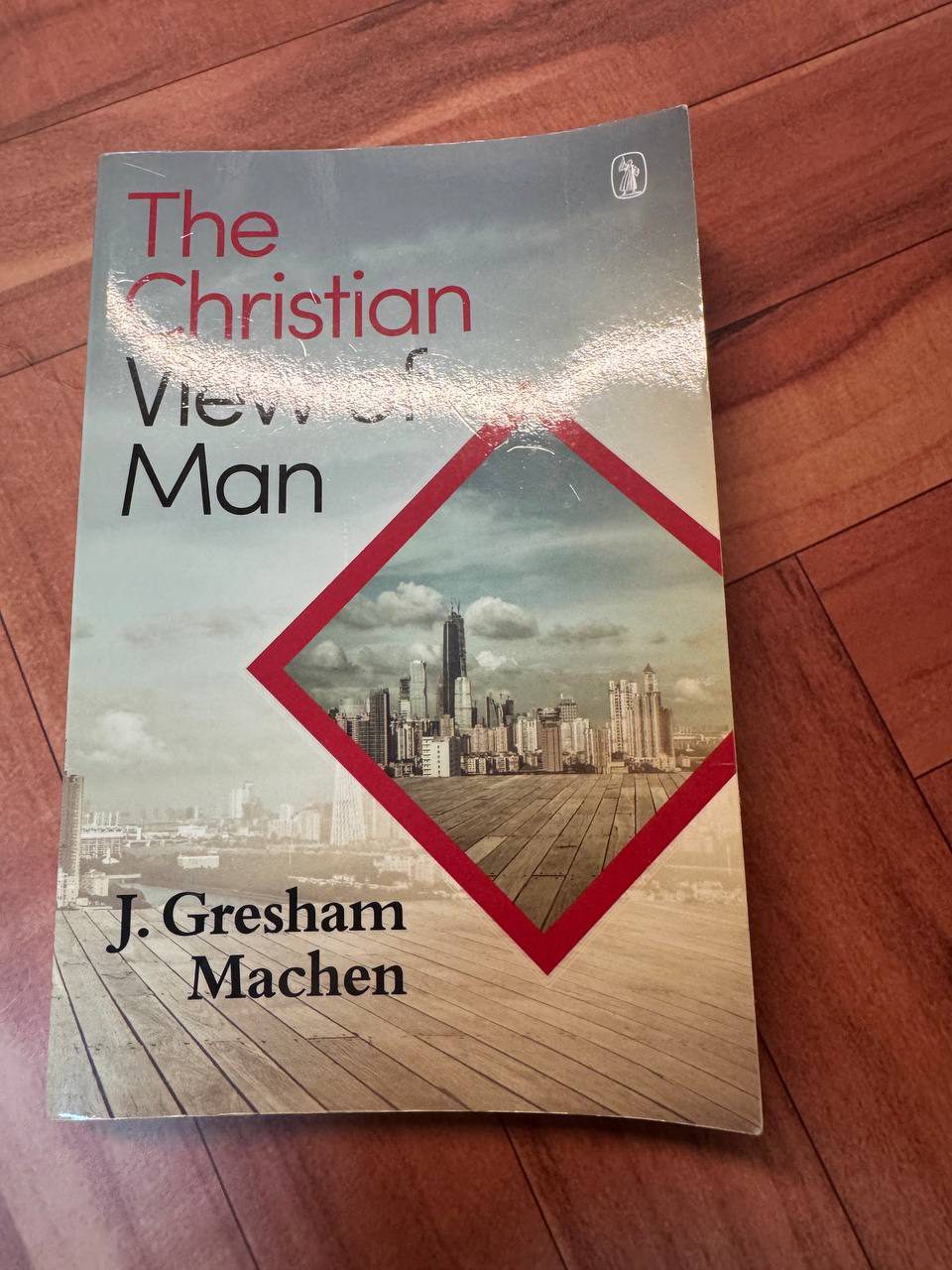 木の床に置かれたJ・グレシャム・メイチェン『The Christian View of Man』のペーパーバック表紙。赤いひし形の中に高層ビル群の写真が配置されている