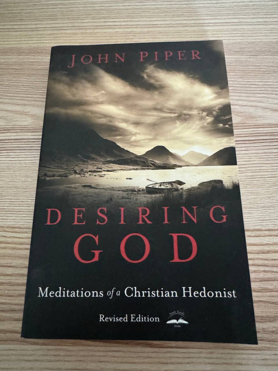 ジョン・パイパー『Desiring God』改訂版の表紙。暗い雲の空と山あいの湖、岸辺に引き上げられた小舟の写真の上に、赤い文字で DESIRING GOD、白で Meditations of a Christian Hedonist と入っている