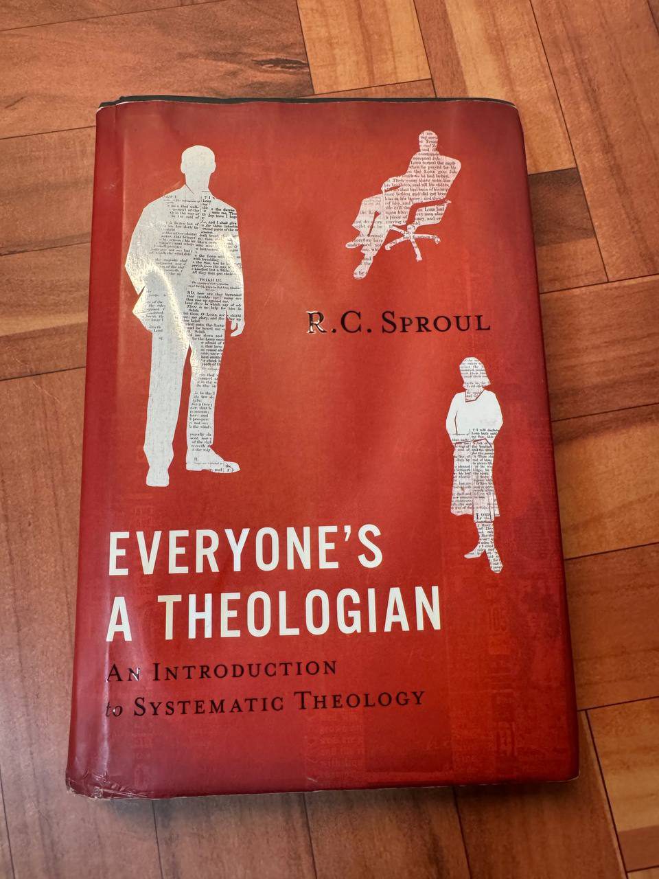 木の床に置かれた赤い表紙のR・C・スプロール『Everyone’s a Theologian』のハードカバー。紙面を切り抜いた人型のシルエットが三つ配置されている