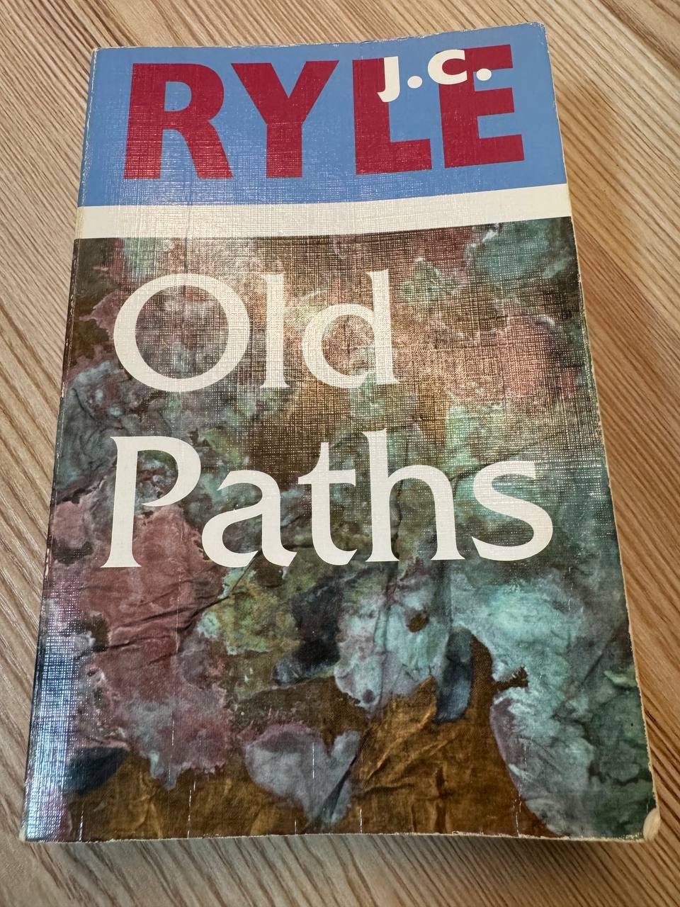 木のテーブルに置かれた J.C. RYLE『Old Paths』のペーパーバック。表紙は青と赤のタイトル帯に、緑と赤茶が混ざった抽象的な絵。角が擦れて白くなっている