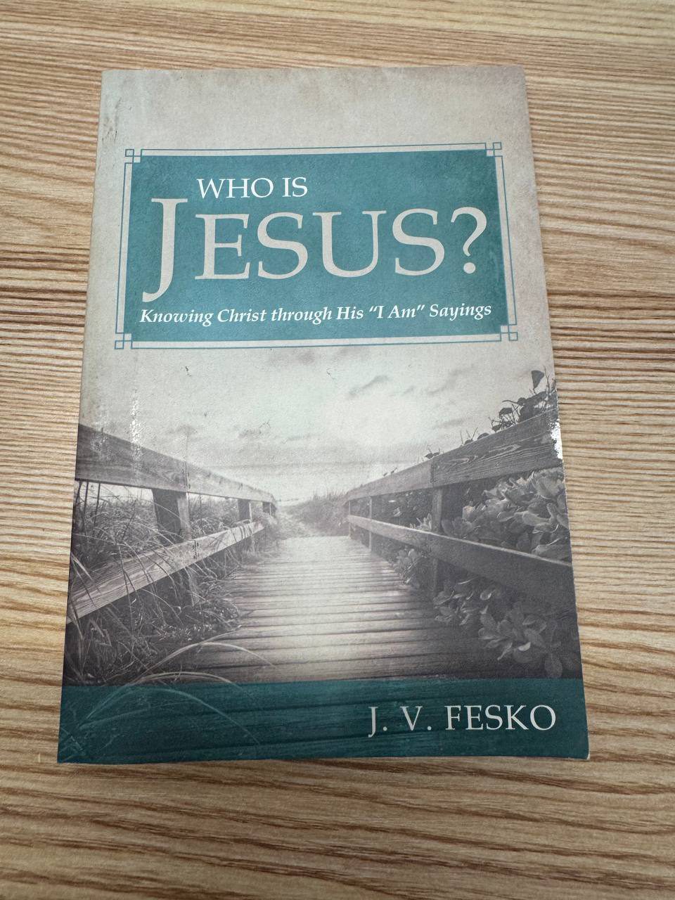 J. V. Fesko『WHO IS JESUS?』の表紙。色あせた紙のような背景に、ティール色の枠と帯が配され、白い大文字で「WHO IS JESUS?」、副題に「Knowing Christ through His ‘I Am’ Sayings」とある。中央には草に囲まれた木の遊歩道のモノクロ写真、下部に著者名 J. V. FESKO が置かれている
